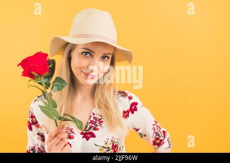 Ein blondie Mädchen trägt einen Hut und hält eine schöne Rose auf einem orangen Hintergrund und schaut auf die Kamera. Hochwertige Fotos Stockfoto