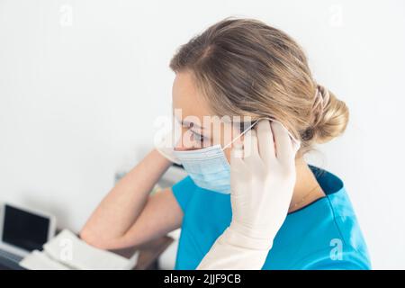 Professionelle Ärztin mit blonden Haaren, die eine Schutzmaske aufsetzt, während sie im Krankenhaus weiße Handschuhe und blaue medizinische Uniform trägt. Hochwertige Fotos Stockfoto