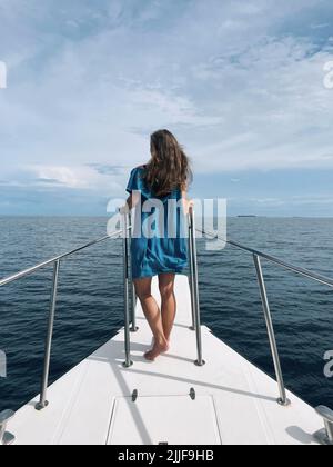 Rückansicht einer jungen Frau in blauem Kleid, die auf einem Bug einer Yacht steht Stockfoto