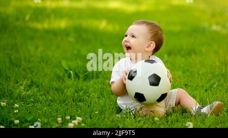 Happy Kleinkind Baby Junge spielt mit einem Fußball auf dem grünen Gras. Lächelndes Kind in weißen Kleidern sitzt mit einem Ball in den Händen, im Alter von einem y Stockfoto