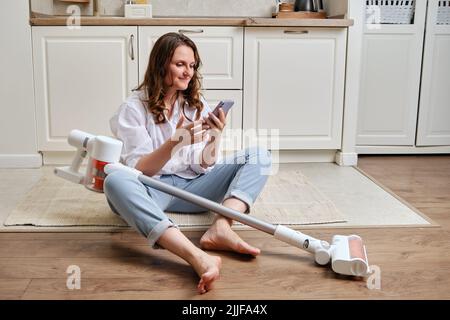 Eine glückliche Frau mit einem Telefon richtet einen tragbaren kabellosen Staubsauger auf dem Boden ein. Lächelnde Frau nach dem Reinigen des Fußbodens in der Wohnung mit Stockfoto