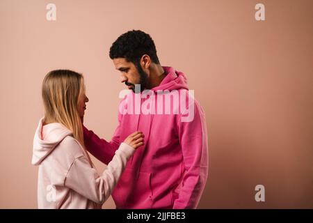 Romantisches Modestudio-Portrait eines jungen Biracial-Paares verliebt in Hoodie posiert auf rosa Hintergrund. Stockfoto