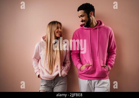 Romantisches Modestudio-Portrait eines jungen Biracial-Paares verliebt in Hoodie posiert auf rosa Hintergrund. Stockfoto