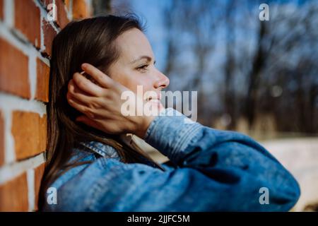 Seitenansicht Porträt einer jungen Frau in Denim-Jacke, die im Sommer die Ziegelwand im Freien lehnt Stockfoto