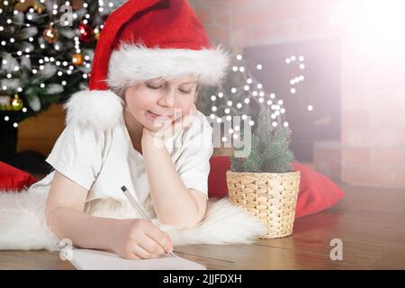 Frohe Weihnachten Kinder. Knabenkind im Weihnachtsmannhut schreibt einen Brief an den Weihnachtsmann unter dem Weihnachtsbaum mit Weihnachtslichtern, in der Nähe des Kamins. Familie Stockfoto