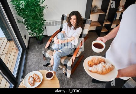 Glückliche Frau, die auf einem Stuhl saß und lächelte, während der Mann eine Tasse Kaffee und Croissants hielt. Verliebte Paare verbringen gemeinsam ihre Freizeit und genießen das Frühstück im gemütlichen Haus. Stockfoto