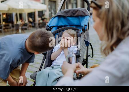 Mutter und kleiner Bruder beruhigen ein weinendes Mädchen, das in einem Kinderwagen sitzt, während sie die Straße entlang gehen Stockfoto