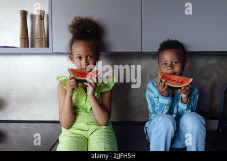 Multirassische Kinder essen Melone in der Küche an heißen sonnigen Tagen. Stockfoto