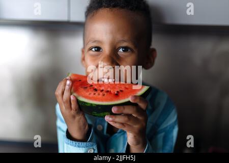 Multirassischer Junge, der an heißen sonnigen Tagen in der Küche Melone isst. Stockfoto