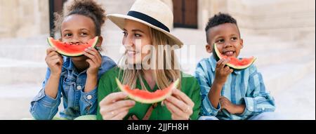 Multirassische Kinder mit Mutter essen Wassermelone auf der Straße an heißen sonnigen Tagen, Sommerurlaub Reisekonzept. Stockfoto