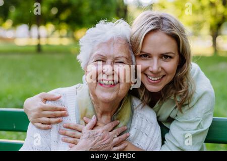 Porträt einer erwachsenen Enkelin, die ihre ältere Großmutter umarmt, als sie auf der Bank im Park sitzt. Stockfoto