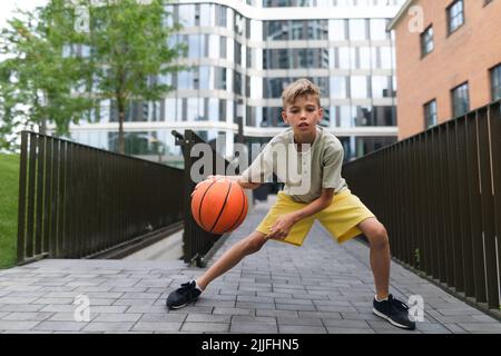 Kaukasischer Junge, der im öffentlichen Stadtpark mit einem Basketballball dribbelt und dabei die Kamera anschaut. Stockfoto