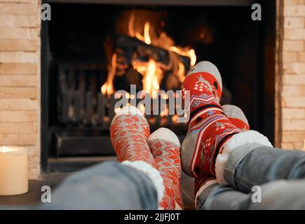 Sich um den Kamin herum zu entspannen. Nahaufnahme eines Paares, das Weihnachtssocken trägt, während es sich mit den Füßen am Kamin zu Hause entspannt. Stockfoto