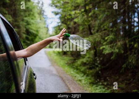 Frauenhand, die Plastikflasche vom Autofenster auf der Straße in grüner Natur, Umweltschutz, Konzept der globalen Erwärmung wegwirft Stockfoto