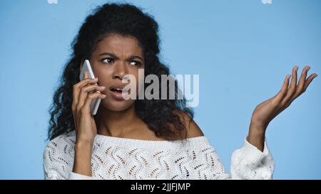 Unzufrieden afroamerikanische Frau in gestricktem Pullover gestikuliert, während sie auf dem Handy isoliert auf blau sprach Stockfoto