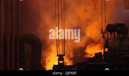 Hintergrund der metallurgischen Schwerindustrie. Große dunkle Werkstatt der Gießerei in Puffs von orangenen Rauch aus dem Hochofen. Stockfoto