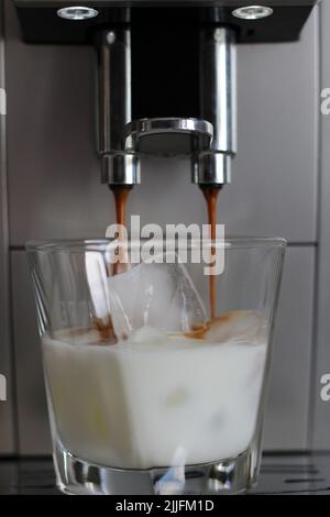 Kaffeemaschine zum Ausgießen von Espresso in Glas mit Milch und Eiswürfeln. Eiskaffee Machen. Erfrischende Getränke Im Sommer. Stockfoto