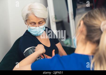 Eine Ärztin in dunkelblauer Uniform, die den Blutdruck einer alten Dame mit einer Gesichtsmaske an einem Blutdruckmessgerät überprüft. Hochwertige Fotos Stockfoto