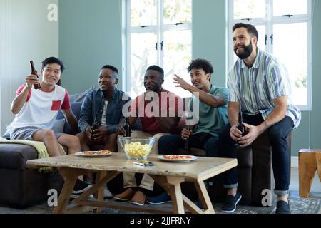 Aufgeregte, multirassische männliche Freunde mit Bierflaschen und Snacks, die sich das Spiel ansehen, während sie sich auf Limonade entspannen Stockfoto