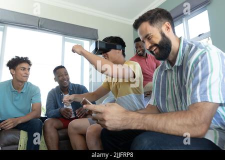 Multirassische Freunde, die lachen und den Mann im Virtual Reality-Simulator betrachten, der zu Hause gestikelt wird Stockfoto