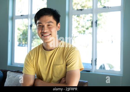 Porträt eines lächelnden chinesischen jungen Mannes mit gekreuzten Armen, der zu Hause gegen die Fenster stand Stockfoto