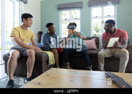 Multirassischer Mann mit digitalem pc und Freund, der zu Hause über ein Virtual-Reality-Headset auf dem Sofa spielt Stockfoto