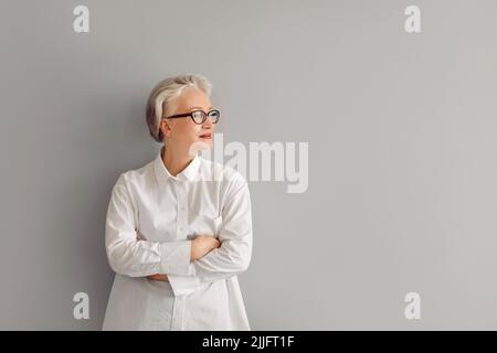 Porträt einer selbstbewussten reifen Geschäftsfrau in Hemd und Brille, die an der grauen Copy Space Wand steht Stockfoto