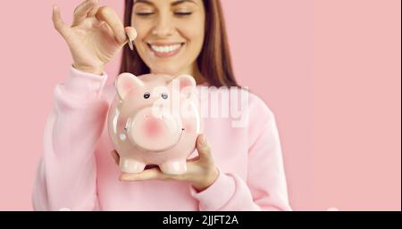 Eine glückliche junge Frau, die etwas Geld spart, steckt eine Münze in ihr rosafarbenes Sparschwein Stockfoto