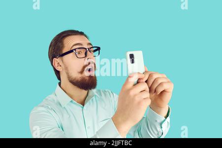 Lustig erstaunt männlich in Brille lesen schockierende Nachrichten auf Handy auf hellblauem Hintergrund. Stockfoto