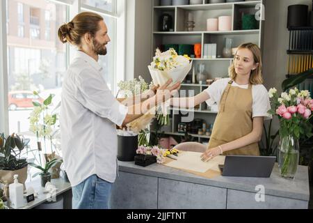 Ein junger bärtiger Mann kauft einen schönen Blumenstrauß für den Urlaub eines Mädchens in einem gemütlichen Blumenladen. Floristik und Blumenstrauß in einem Blumenladen. Kleinunternehmen. Stockfoto