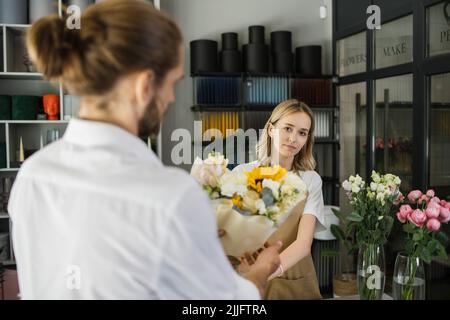 Ein junger bärtiger Mann kauft einen schönen Blumenstrauß für den Urlaub eines Mädchens in einem gemütlichen Blumenladen. Floristik und Blumenstrauß in einem Blumenladen. Kleinunternehmen. Stockfoto