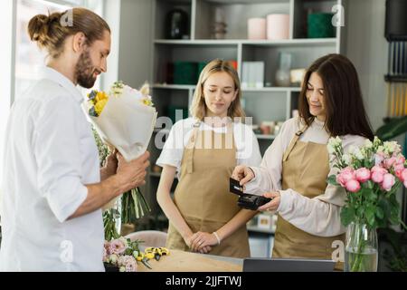 Porträtaufnahme von zwei jungen charmanten Blumenhändlern, die einem attraktiven bärtigen männlichen Kunden, der per Kreditkarte bezahlt, einen Strauß geben. Stockfoto