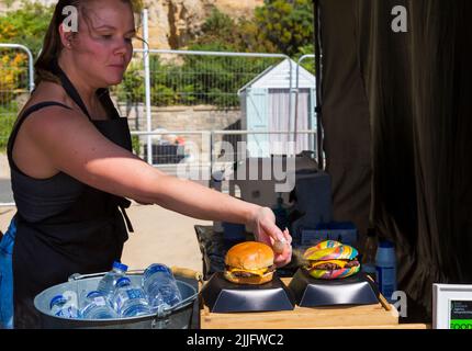 Unicorn Burger Hampshire Hacksteak Regenbogen Bagel Glitzer zum Verkauf auf Food Stand bei der Veranstaltung in Poole, Dorset UK im Juli - Frau sprüht Glitzer auf Stockfoto