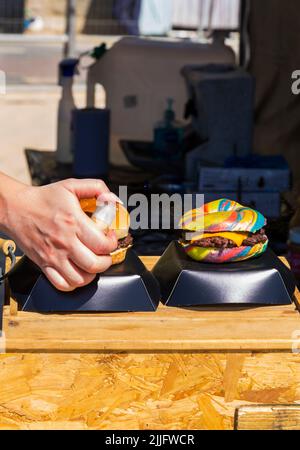 Unicorn Burger Hampshire Hacksteak Regenbogen Bagel Glitzer zum Verkauf auf Food Stand bei der Veranstaltung in Poole, Dorset UK im Juli - Frau sprüht Glitzer auf Stockfoto