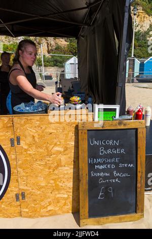 Unicorn Burger Hampshire Hacksteak Regenbogen Bagel Glitzer zum Verkauf £9 auf Food Stand bei der Veranstaltung in Poole, Dorset UK im Juli - Frau sprüht Glitzer auf Stockfoto