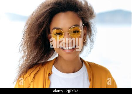 Nahaufnahme einer schönen, fröhlichen, trendigen afroamerikanischen Frau mit lockigem Haar, mit orangefarbener Brille, weißem T-Shirt und orangefarbenem Hemd, steht im Freien, blickt auf die Kamera und lächelt freudig Stockfoto