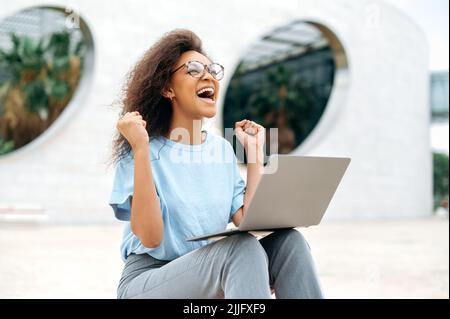 Fröhlich aufgeregt afroamerikanische junge Frau mit Brille, nutzt Laptop, während sie im Freien sitzt, die Hände vor Aufregung hebt, Fäuste streicht, Erfolg feiert, gewinnt, glücklich lächelt. Gute Nachrichten, gewinnen Stockfoto