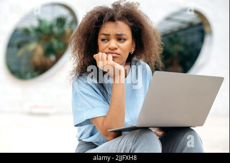 Frustrierte afroamerikanische junge Frau mit Laptop. Gemischte Rasse weibliche Geschäftsfrau, Freiberuflerin oder Studentin, sitzt im Freien mit Laptop, schaut weg, traurig wegen der schlechten Nachrichten, müde von langweiliger Arbeit Stockfoto