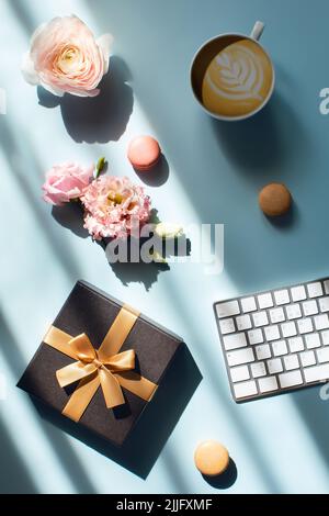 Freiberuflicher Ferienarbeitsplatz. Blauer Tisch am Fenster mit Cappuccino, Kuchen, Geschenk und Blumen. Stockfoto