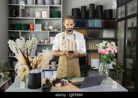 Kleinunternehmen. Junger bärtiger männlicher Florist im Blumenladen, der eine Nachricht an den Kunden mit einem Smartphone schreibt. Blumendesign Studio, Dekorationen und Arrangements. Blumen Lieferung, Schöpfung Auftrag. Stockfoto