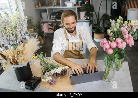 Hübscher Blumenladen, männlicher Besitzer, der Schürze trägt und im Geschäft arbeitet. Attraktive junge Floristin mit Laptop, um mit Kunden, Kunden zu chatten, um in den Neuigkeiten zu blättern. Social Media, Technologie-Geschäftskonzept Stockfoto