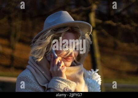 Eine Frau in einem Hut mit einem flatternden Haarsträhnen telefoniert in einem Herbstpark bei Sonnenuntergang. Sie ist glücklich und lächelt. Weicher Fokus. Spot. Ein geldautomat Stockfoto