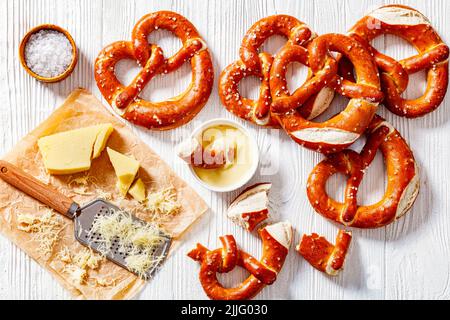 Weiche Brezeln in Form von Knoten gebacken und mit Salz auf weißem Holztisch mit Käsesauce und geriebenem Käse bestreut, horizontale Ansicht von oben, Stockfoto