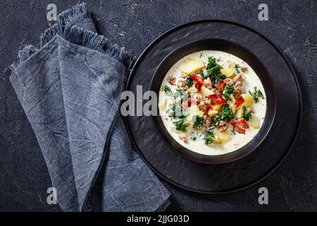 Reiche, cremige toskanische Suppe, Zuppa Toscana beladen mit italienischer Wurst, Grünkohl, gebratenem Speck und Kartoffeln in schwarzer Schüssel auf grauem Betontisch, horizontal Stockfoto