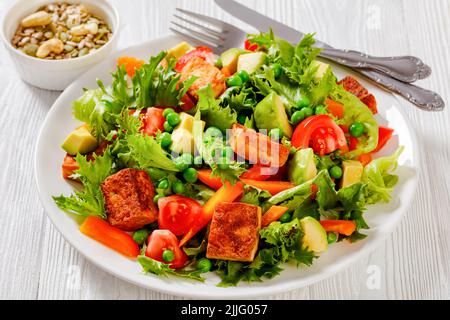 Tofu-Salat mit Avocado, knackigen Salatblättern, grünen Erbsen, Tomaten, gelber Paprika auf weißem Teller, horizontale Ansicht von oben Stockfoto