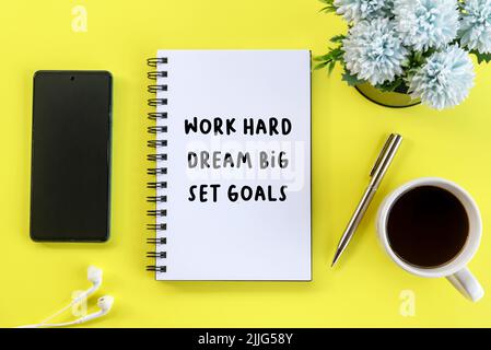 Arbeiten Sie hart, träumen Sie sich Ziele. Motivierendes Zitat in flachem Lay-Geschäftskonzept auf Notebook mit Stift, Telefon, Kaffee, Blumen auf gelben Tischhintergründen. Stockfoto