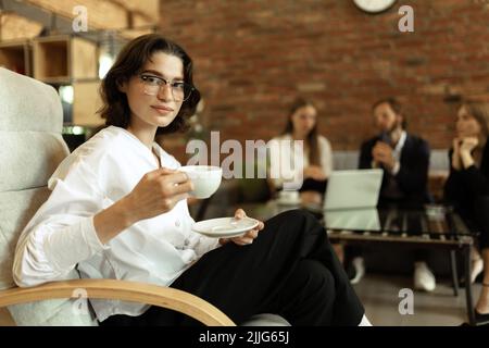 Junge Frau in offizieller Kleidung und stilvollen Gläsern, Kaffee trinken, Pause. Weibliche Geschäftsmannes, die mit dem Team im Büro arbeitet Stockfoto