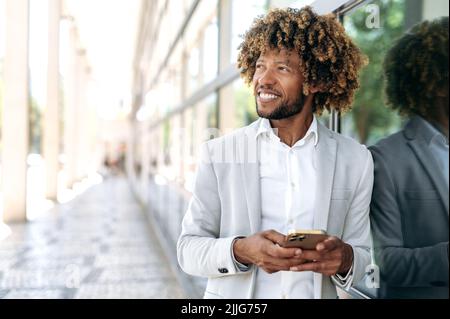 Ein gutaussehender, glücklicher brasilianischer oder afroamerikanischer Mann mit lockigen Haaren, elegant gekleidet, im Freien in der Nähe des Geschäftszentrums stehend, Mobiltelefon verwendend, wegblickend, träumend, denkend, Lächelnd Stockfoto