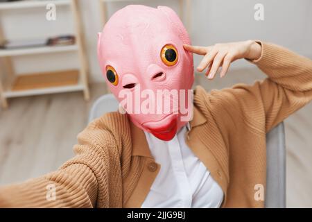 Lustige blonde Dame in rosa Fischmaske warmen Pullover tun Selfie verwenden Telefon sitzen im Sessel im modernen Zuhause Interieur. Pause von der Arbeit, Pause machen Stockfoto