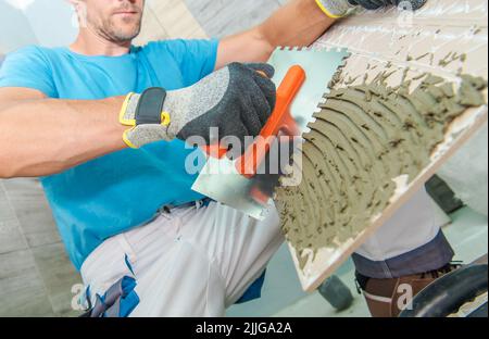 Kaukasischer Arbeiter, der vorsichtig mit Spezialwerkzeug Klebstoff auf die Fliese legt. Thema Renovierung und Renovierung des Badezimmers. Stockfoto